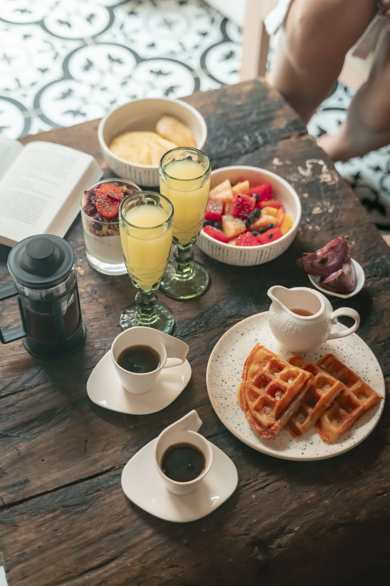 Desayuno servido sobre mesa de madera: waffles, café, jugo de naranja y fruta fresca, sobre piso de baldosa hidráulica.
