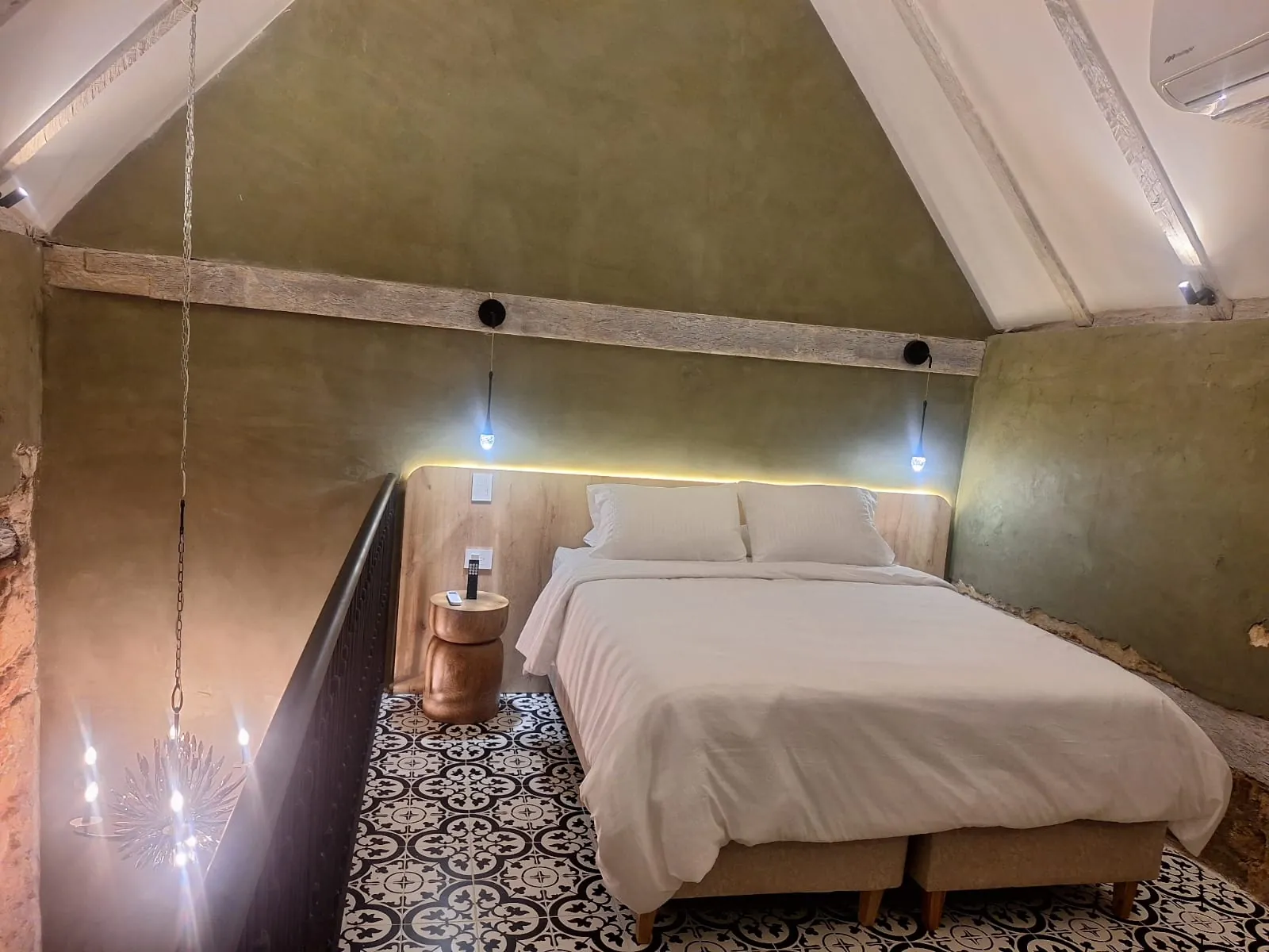 Habitación con muro en verde oliva, cama tendida en blanco y mezzanine de madera oscura.